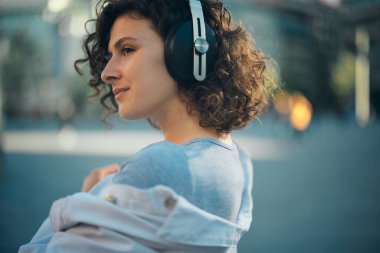 Kıvırcık saçlı bir kızın portresi. Rastgele giyinmiş, dışarıda dikilip başka tarafa bakıyor..