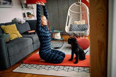 Sakin, esnek, son sınıf öğrencisi bir kadın evde yoga minderinde diz çöküp köpeğiyle yoga yapıyor..