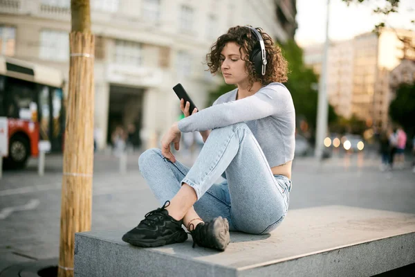 Młoda Dziewczyna Kręconymi Włosami Siedzi Zewnątrz Słucha Muzyki Przez Telefon — Zdjęcie stockowe