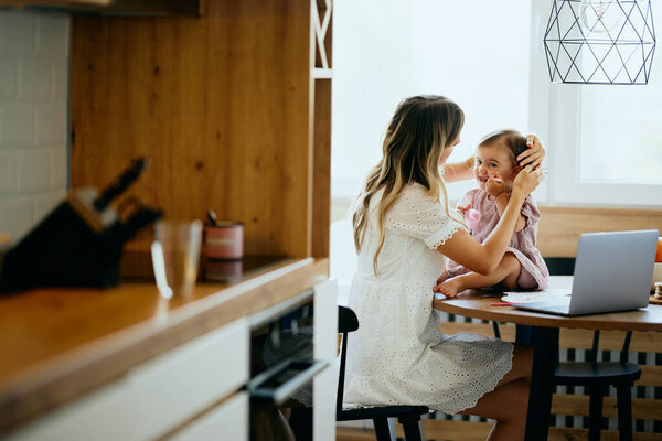 A young mother sitting at the dining table at home and adjusting hair clips on her daughter's hair. A baby girl sitting on a table and chewing a spoon.