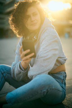 Kıvırcık saçlı genç bir kadın sokakta oturuyor ve mesaj atıyor. Boynunda kulaklık var..