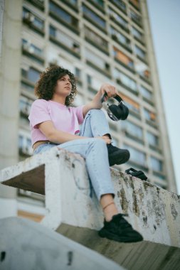 Kıvırcık saçlı, açık havada oturan ve elinde kulaklık tutan çekici bir genç kadının düşük açılı görüntüsü. Kentsel yaşam kavramı.