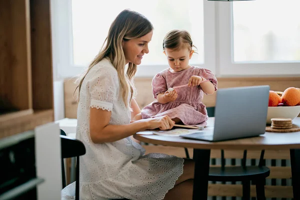 Stay at mother home sitting in kitchen and getting work done while her baby girl helping her.