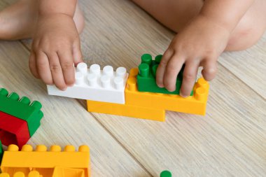 Küçük çocuk yerde plastik renkli oyuncak bloklarıyla oynuyor. Evde renkli plastik tuğlalarla oynayan çocukların ellerini kapayın. Yeni yürümeye başlayan çocuk parlak tuğlalardan eğleniyor ve inşa ediyor..