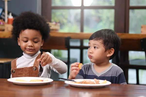 Cute African American boy with curly and adorable Asian kid eating meal at the table indoor, happy children having food in a restaurant, enjoy their meal together.