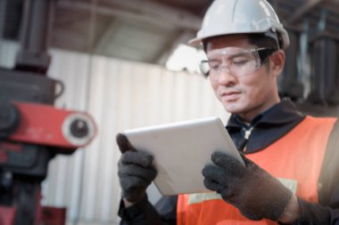 Kasklı ve güvenli gözlükler takan endüstri mühendisi, fabrika fabrikasındaki işletim makineleri için dijital tablet tutuyor, endüstri konseptinde makine ile çalışıyor.