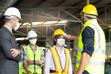 Foreman dijital termometreye sahip, yüz maskesi takan ekran işçileri için sıcaklık ölçer, Covid-19 salgınını önler, endüstriyel mühendisler imalat fabrikasında sırada..