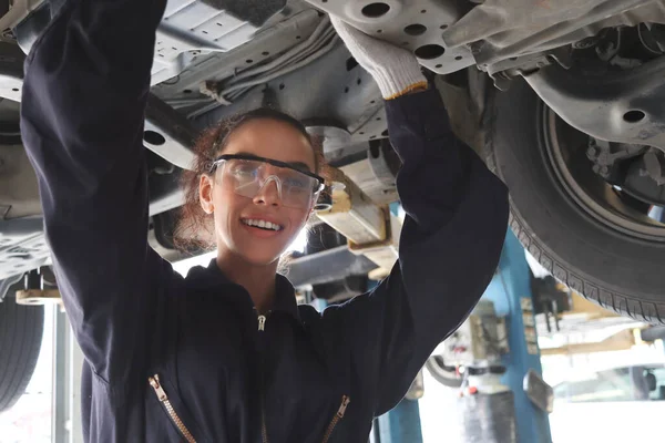 Female auto mechanic working in garage, car service technician woman checking and repairing the customer car at automobile service center, inspecting car under body and suspension system, vehicle repair service shop concept.