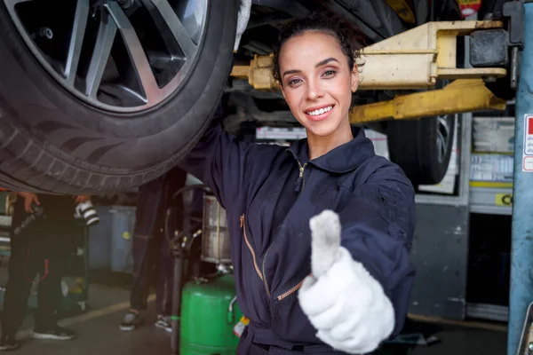 Female auto mechanic work in garage, car service technician woman give thumb up, repair customer car at automobile service center, inspecting car under body and suspension system, vehicle repair service shop.