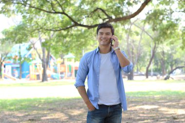 Bahçe parkında akıllı telefon kullanan sıradan giyinmiş, cep telefonuyla konuşan genç yakışıklı bir adam.