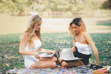 Öğrenci kampüs parkında ders çalışıyor, kitap okuyor ve dizüstü bilgisayar kullanıyor, parkta piknikte mutlu arkadaşlar, bahçede birlikte vakit geçirirken eğlenen iki genç kız, açık havada arkadaşlık kavramı.