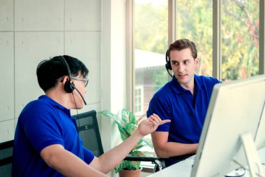 Man with headphones working at call center service desk consultant with his teammates, helping and training his colleague to talking with the customer on hands-free phone, happy workplace concept clipart