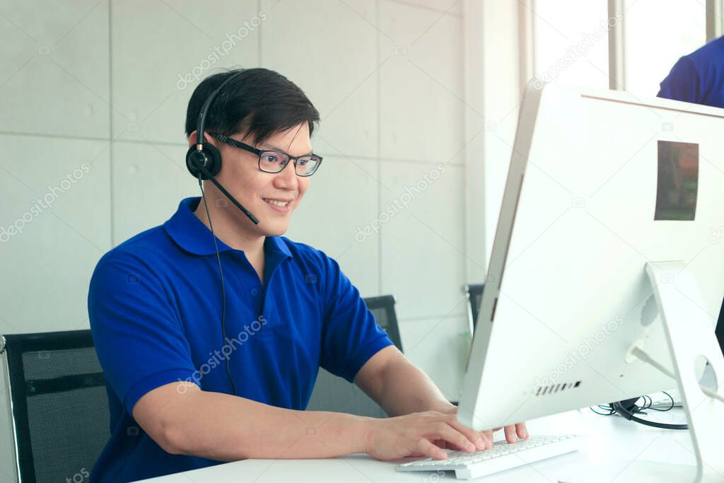 Happy smiling Asian man with headphones working at call center service desk consultant, talking with the customer on hands-free phone, happy workplace concept 