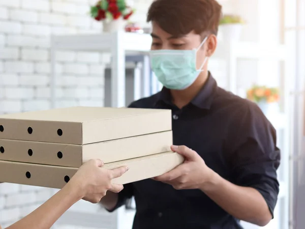 Hands deliver goods to the recipient customer. Young man wearing face mask receiving delivery to pizza box to prevent covid19 infection, social distancing and new normal shipping and courier delivery services express concept.