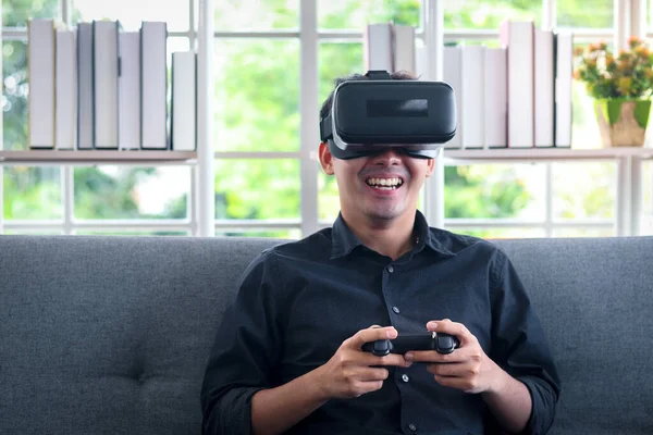 Asian young happy man wearing virtual reality device (VR glasses) for playing the video game while sitting on sofa in his apartment, cheerful male having fun with new trends technology, technologies of the future concept.