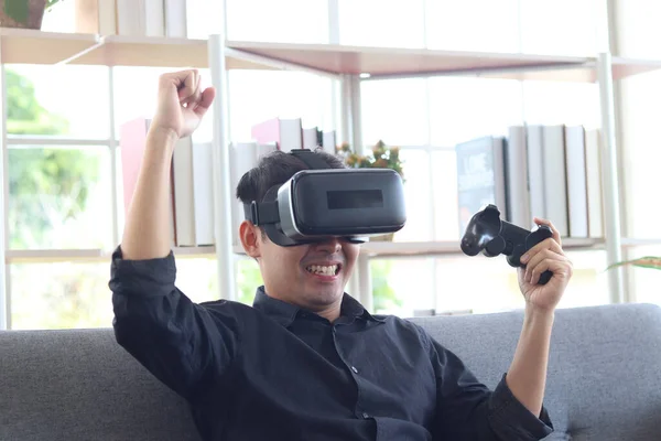 Asian young happy man wearing virtual reality device (VR glasses) for playing the video game while sitting on sofa in his apartment, cheerful male having fun with new trends technology, technologies of the future concept.