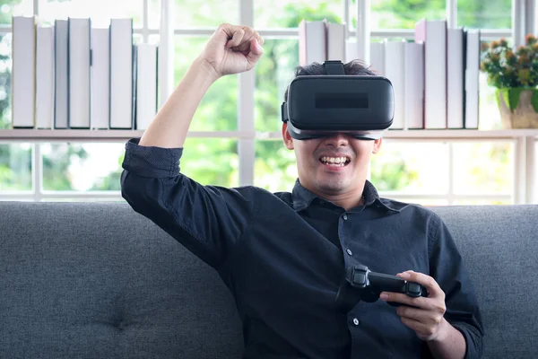 Asian young happy man wearing virtual reality device (VR glasses) for playing the video game while sitting on sofa in his apartment, cheerful male having fun with new trends technology, technologies of the future concept.