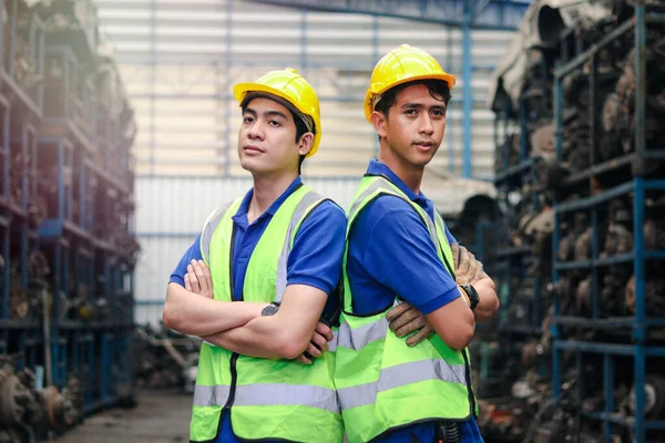 Portrait of two industrial engineer workers man wearing helmet with arms crossed, standing at manufacturing plant factory with many engine parts as blurred background