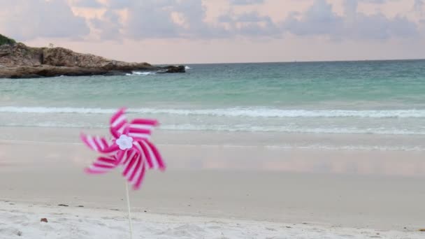 粉红砂轮玩具在白色沙滩上旋转 早上海滩上有两个五彩斑斓的风力涡轮机 背景是夏天的大海和橙色日出的天空 — 图库视频影像