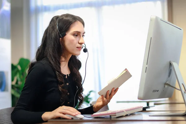 Young beautiful female with headphones working at call center service desk consultant, talking with the customer on hands-free phone, woman customer support agent with headset and desktop computer working at call center.