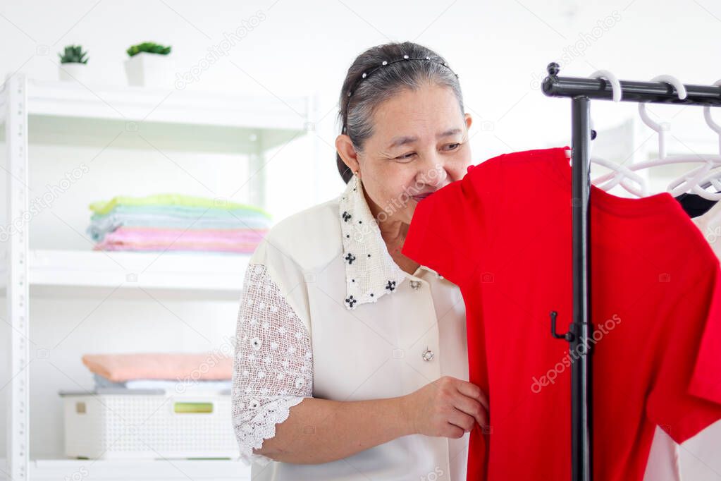 Happy smiling senior elderly woman housewife smelling T-shirt, sniffing from clothes, hanging clothes indoor to dry, older Asian female doing laundry at laundry room, dried laundry, grandma doing housework and cleaning clothes at house.