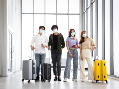 Koronavirüs enfeksiyonunu önlemek için maske takan Asyalı genç turist grubu havaalanında duruyor, havaalanında havayolu uçuşunu bekliyor, sosyal uzaklık ve yeni normal seyahat kavramı.