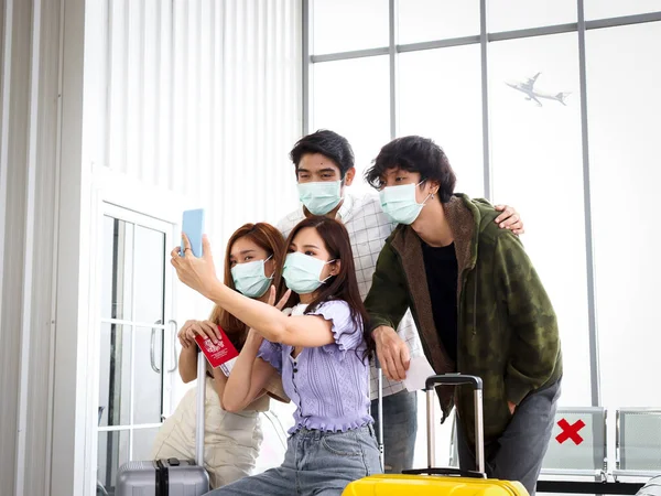 Asian young tourist friend group wearing face mask to prevent coronavirus infection using mobile phone for taking selfie photos during waiting airline flight at airport terminal, social distancing and new normal travel concept.