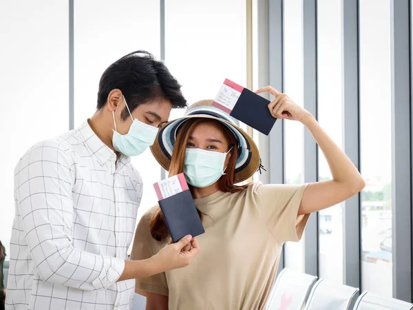 Asian young tourist lover couple wearing face mask to prevent coronavirus infection, holding passport and boarding pass (mock up), waiting airline flight at airport terminal, social distancing and new normal travel concept.