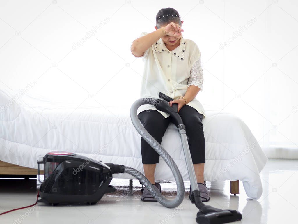 Asian senior elderly woman housewife vacuuming the floor at bedroom with vacuum cleaner, feeling tired and exhausted, resting by sitting in bed, grandma doing housework at house.