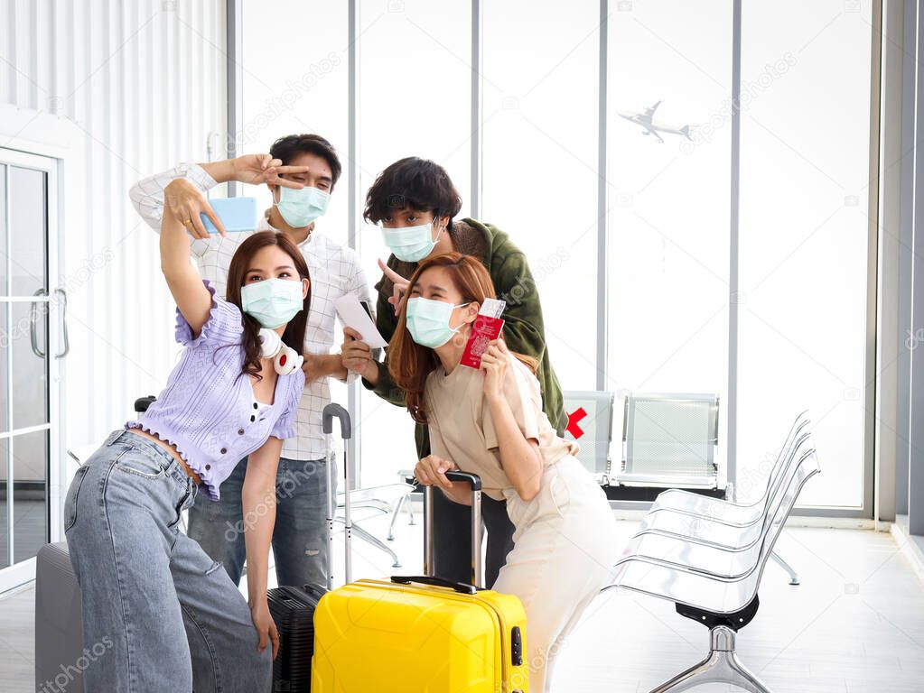 Asian young tourist friend group wearing face mask to prevent coronavirus infection using mobile phone for taking selfie photos during waiting airline flight at airport terminal, social distancing and new normal travel concept.
