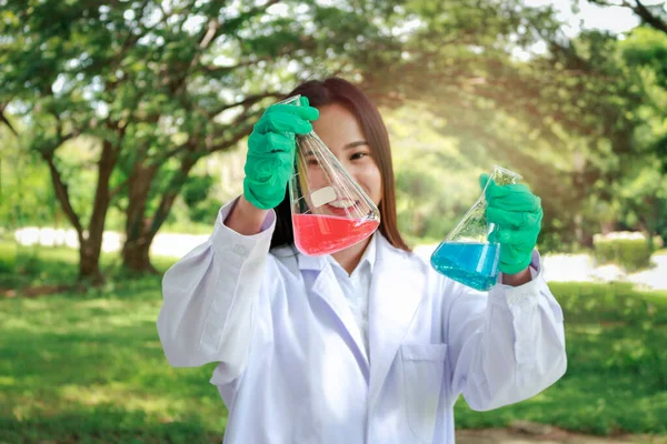 Scientist doing science experiment surround green nature park garden, young beautiful researcher woman holding conical flask with chemical solutions, green chemical and environmental friendly science concept