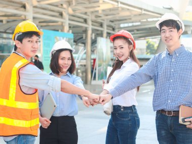 Şehir merkezindeki ofisin dışında el ele tutuşan, güvenlik yeleği ve kask giyen dört genç Asyalı erkek ve kadın mühendis grubu, güçlü bir takım çalışması, birlik ve takım çalışması olarak el ele tutuşan ortaklar.