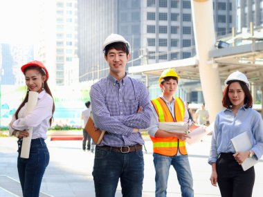 Asyalı dört zeki genç erkek ve kadın mühendis grubunun portresi, güvenlik yeleği ve miğferi giyen personel işçi takımı, şehir merkezindeki ofisin dışında duran gökdelen binası bulanık arka plan olarak görülüyor..