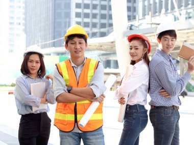 Asyalı dört zeki genç erkek ve kadın mühendis grubunun portresi, güvenlik yeleği ve miğferi giyen personel işçi takımı, şehir merkezindeki ofisin dışında kolları çapraz duran, gökdelen binası olarak bulanık arka plan.