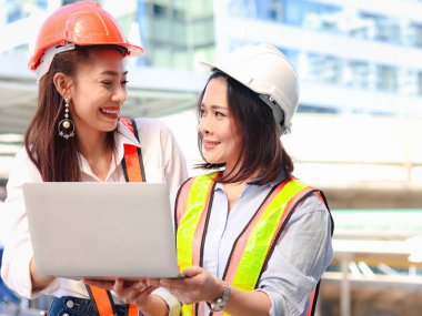 İki güzel Asyalı mühendis kadın güvenlik yeleği ve kask giyiyorlar, birlikte dizüstü bilgisayar kullanıyorlar, güzel kadınlar şehir merkezindeki ofisin önünde yürürken proje çalışmaları hakkında tartışıyorlar.. 