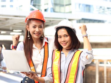 Güvenlik yeleği ve kask giyen iki güzel Asyalı mühendis kadın proje hakkında tartışmak için dizüstü bilgisayar kullanıyorlar. Şehir merkezindeki ofisin dışında yürürken neşeli genç kadınlar el kaldırıyor.. 