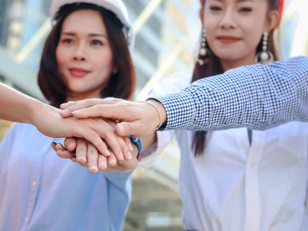 Group of four young Asian engineers wearing safety helmet, putting hands join together at outside office in downtown city, partnership colleagues holding hands as commitment of strong teamwork, unity and teamwork join hands support concept.