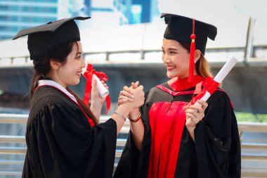 Mutlu gülümseyen iki mezun öğrencinin portresi, genç ve güzel Asyalı kadınların birbirlerine güçlü bir şekilde el ele tutuşmaları, mezuniyet günlerinde çok gururlu olmaları, mezuniyet günlerinde başarılı eğitimlerini kutlamaları,
