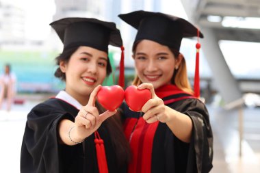 Elinde küçük kırmızı kalp hediyeleri olan ve kameraya hediye veren mutlu gülümseyen iki mezun öğrenci, mezuniyet gününde başarılı bir eğitimi kutlayan genç ve güzel Asyalı kadınlar, üniversite ya da üniversite eğitimini bitiren insanlar.