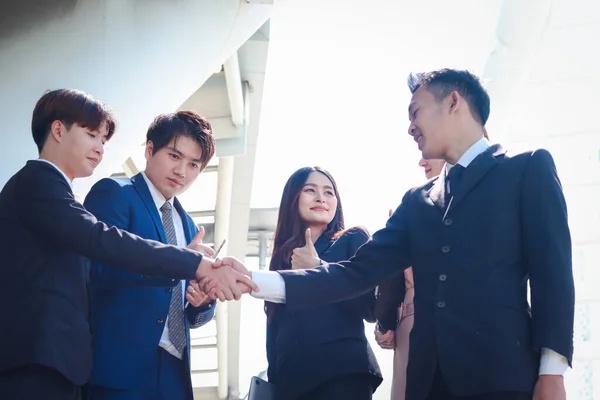 Group of young Asian achievement successful businesspeople shaking hands after sign contract, corporate group giving thumbs up and appreciating the good result, businessman and woman have meeting at outdoor in city.