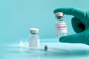 Mavi arka plan ve fotokopi alanı üzerinde bulanık iğne şırıngası, koronavirüsün önlenmesi için aşı ve bağışıklık sistemi (COVID-19) ile iğne tedavisinde kullanılan sıvı tıbbi aşı içeren cam şişeyi tutan eldiven.