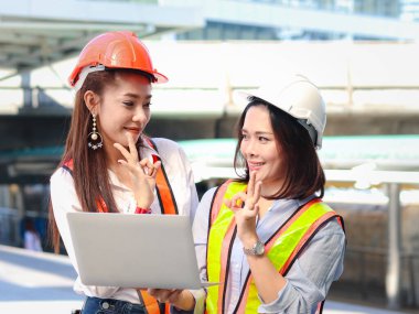 İki güzel Asyalı mühendis kadın güvenlik yeleği ve kask giyiyorlar, dizüstü bilgisayar kullanıyorlar, kadınlar proje hakkında tartışıyorlar ve şehir merkezindeki ofisin dışında yürürken el işareti gösteriyorlar.. 