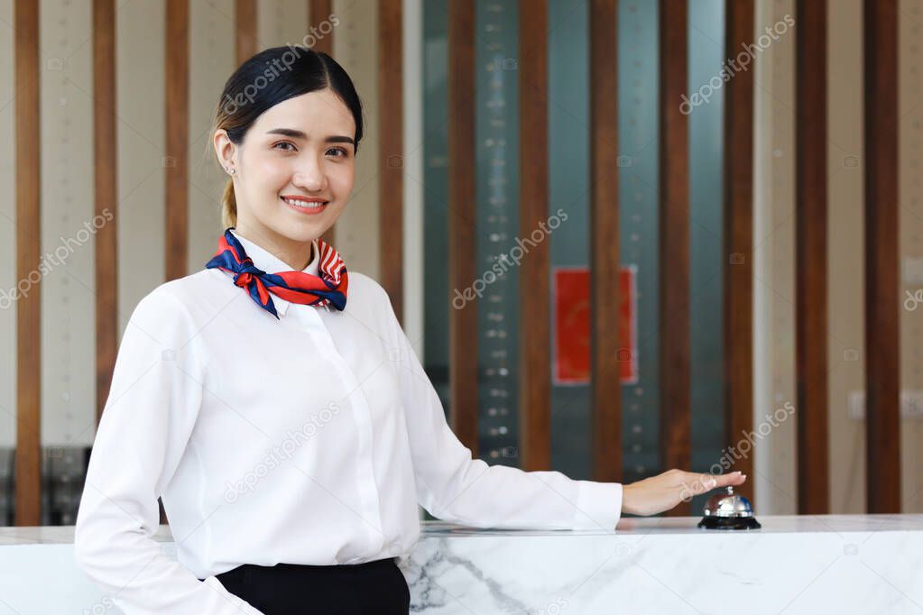 Portrait of smiling young beautiful Asian female receptionist reaching hand to ring silver bell on hotel reception service desk, check in hotel service on vacation concept.