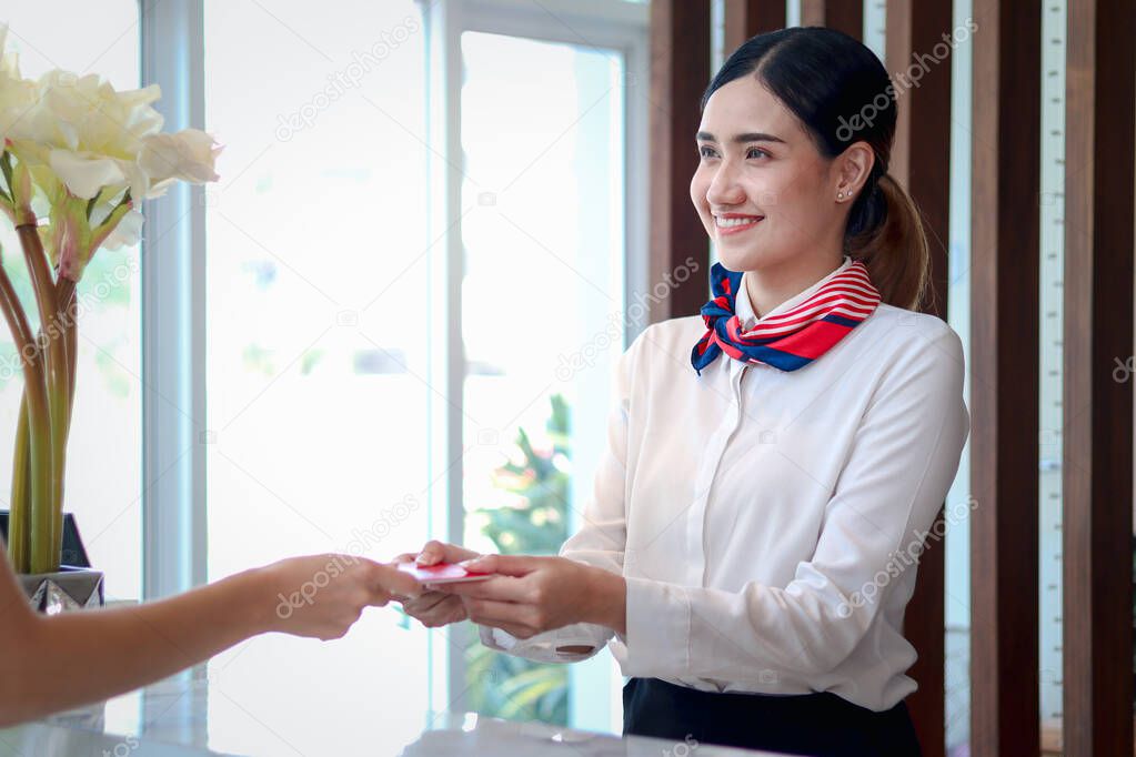 Smiling young beautiful Asian female receptionist giving key card to customer hand at hotel reception counter desk, check in hotel service on vacation concept. 