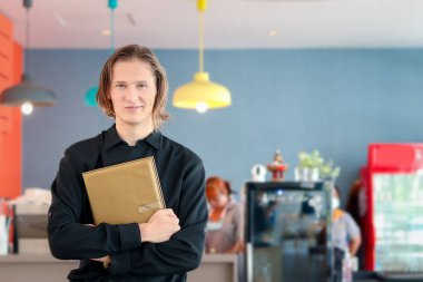 Siyah üniformalı, gülümseyen, yakışıklı erkek garsonun portresi. Kollarını kavuşturup kafe veya restoranda menüyü tutuyor.