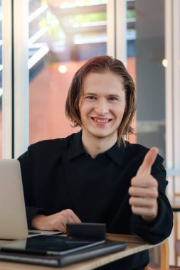 Masada dizüstü bilgisayarla oturup çalışırken kameraya baş parmağını veren mutlu genç adam..