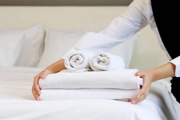 Room service maid cleaning and making bed hotel room concept, woman hands putting stack of fresh white bath towels on bed sheet.