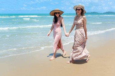 Büyük şapkalı ve güneş gözlüklü iki güzel Asyalı kadın arkadaşlarıyla tropikal kumsal mavi denizinde vakit geçirmeyi seviyorlar, yürüyüşe çıkıyorlar, yaz tatilinde dinleniyorlar ve dinleniyorlar.. 