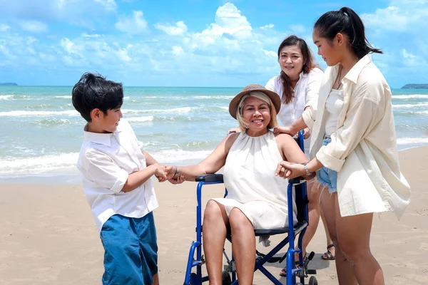 Happy disabled senior elderly woman in wheelchair spending time together with her family on tropical beach. Asian grandma, daughter and grandchildren resting and relaxing on summer holiday vacation.