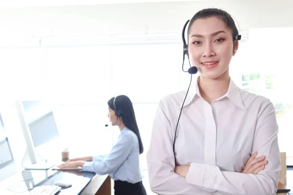 Portrait of smiling beautiful Asian woman with headphones work at call center service desk consultant, call center operator agent at workplace with hands-free phone ready to help customer on hotline.